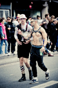 Two individuals walking in a crowded street during a festival or parade. One is shirtless wearing harness gear and sports pants, holding a bottle. The other wears a white bucket hat, harness, and knee-high socks with shorts. Both appear engaged in conversation.