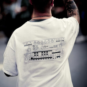 A person wearing a white t-shirt with a detailed illustration of a Roland TB-303 Bass Line synthesizer printed on the back. The person has a tattooed arm raised and is facing away from the camera in a blurred background.