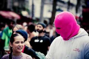 A person wearing a pink ski mask and a white hoodie with text stands next to a smiling woman in a crowd. The background is blurred, showing several other people and buildings, suggesting an outdoor event or protest.