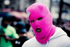 A person wearing a bright pink knitted balaclava stands in an outdoor setting. The mask covers their entire head, with openings for their eyes and mouth. The background is blurred, showing people and buildings. The person is also dressed in a light-colored hoodie.