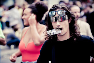 20230923-5H1A9049_MOD_FRAME_resize A person with long hair wears mirrored sunglasses and a futuristic visor covering their eyes. They have two cigarette packs in their mouth. The background is blurred, showing another person with a ponytail wearing an orange top. The scene appears to be outdoors.