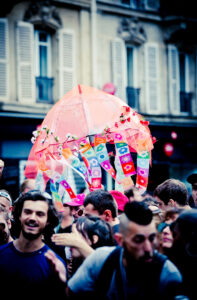 A crowd of people is gathered outdoors in an urban area. In the center, a clear umbrella decorated with colorful ribbons and floral designs is held up. The background features a building with shuttered windows. The atmosphere appears festive.