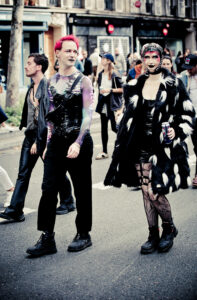 Two individuals dressed in vibrant, alternative fashion walk down a city street. The person on the left wears a colorful, tight outfit with pink hair, and the one on the right in a black-and-white coat, patterned tights, and a hat. Other pedestrians are in the background.