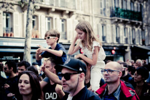 A crowd of people at an outdoor event, including a man in a cap and sunglasses carrying a girl on his shoulders, and another man in a shirt labeled "DAD" carrying a boy on his shoulders. The children are watching attentively. Buildings with shutters are in the background.