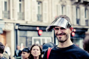 A man in a black shirt smiles while wearing a shiny metallic visor on his head. He has an earring in his left ear and carries a red strap on his right shoulder. The background shows an urban setting with buildings and several other people.