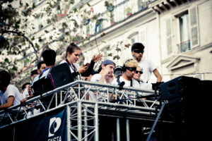 A group of people on a platform, some wearing sunglasses and white shirts, participate in an outdoor event. The setting is urban, with a railing and residential buildings in the background. One person uses a camera, capturing the moment.