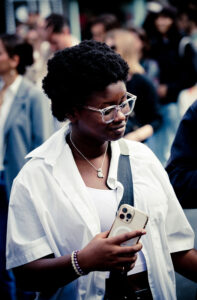 A person with short curly hair and glasses is looking at their phone. They are wearing a white shirt over a white top, a necklace, and a bracelet. In the background, several people are gathered, blurred out.