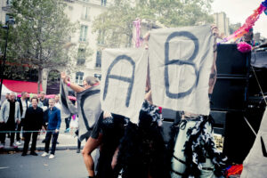 Individuals hold large cloth banners with the letters "A" and "B" at an outdoor event. People in the background watch, with buildings and trees visible. The atmosphere seems festive with colorful decorations.
