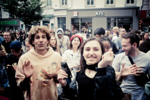A lively crowd of people is gathered outdoors. The foreground shows a man with curly hair and a woman smiling and holding up an object. The background includes several people, a pharmacy, and a KFC restaurant. Everyone appears happy and engaged.