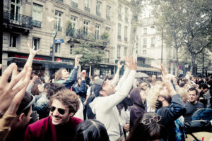 A large crowd of people joyfully gathered on a city street, raising their arms and cheering. The backdrop consists of buildings with shops and balconies. Trees can be seen lining the street, and the overall atmosphere appears lively and celebratory.
