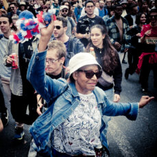 20230923-DSCF0527_MOD_FRAME_resize An older woman in sunglasses, a white hat, and a colorful outfit energetically raises her hand holding pom-poms. She's surrounded by a diverse group of people enjoying a lively street event. Some are taking photos, while others cheer and hold drinks.