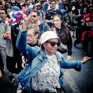 An older woman in sunglasses, a white hat, and a colorful outfit energetically raises her hand holding pom-poms. She's surrounded by a diverse group of people enjoying a lively street event. Some are taking photos, while others cheer and hold drinks.