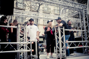A group of people, mostly in casual attire and wearing caps, gather on a metal stage structure. Some are conversing, while a few hold drinks and mobile phones. The background features a cityscape with building facades. It appears to be an outdoor event.