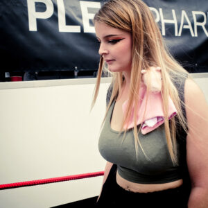 A young woman with long blonde hair, wearing a green top and black pants, has a small plush toy pig draped over her shoulder. She stands outdoors in front of a sign with partial text and a red rope barrier.