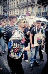 A group of people are walking down a city street during what appears to be a protest or public event. One person prominently wears a colorful, graffiti-style jacket, white-framed sunglasses, and a panda hat. The background shows buildings and trees.
