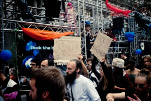 A group of people marching in a protest, holding signs with messages. One sign reads "TOKIO HOTEL," and another says "FUCK LE PATRIARCAT." The crowd is diverse, filled with individuals of varying ages and styles, set against a backdrop of metal scaffolding and banners.