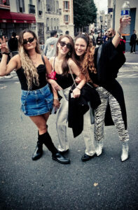 Four women stand on a street, posing cheerfully for the photo. They are dressed in trendy outfits, including jeans, skirts, and stylish boots. One holds up a water bottle while they all smile at the camera. The background shows more people and buildings.