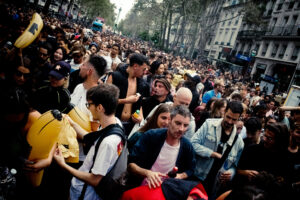 A large crowd of people gathers in the street for a public event. The diverse group includes individuals of various ages, holding inflatable objects, cellphones, and drinks. The setting appears to be an urban area with tall buildings lining the street in the background.