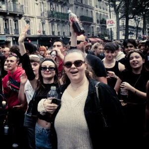 A large group of people are gathered outdoors in a city, enjoying what appears to be a street festival or parade. The crowd appears joyful and engaged, with several individuals raising their hands and holding up bottles. Buildings and trees are visible in the background.