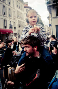 A man carrying a young child on his shoulders in a busy street scene. The child is wearing headphones and a striped shirt while holding a small item. The man, with earplugs in his ears, looks forward amidst a crowd of people and surrounding buildings.