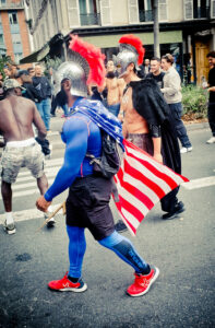 People in a street parade, with two prominently dressed in Spartan-like helmets and red-crested plumes, one wearing a blue outfit with an American flag cape. Others are casually dressed, observing or participating. The event appears festive and lively.