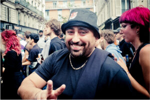 A bearded man wearing a black bucket hat and chain necklace smiles and flashes a peace sign at an outdoor gathering. He is surrounded by people with colorful hairstyles and streetwear attire. In the background, buildings and other event-goers can be seen.