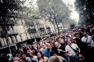 A large crowd of people walking down a street lined with trees and buildings, under a cloudy sky. The street is busy with a diverse mix of individuals, some with raised hands or holding signs, indicating a possible protest or large public gathering.