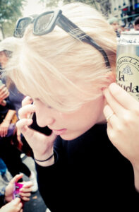 A person with light blonde hair, wearing black sunglasses on their head, holds a black smartphone to their ear with one hand and a can of "La BoudA Blonde" beverage to their other ear amidst a crowd of people outdoors.