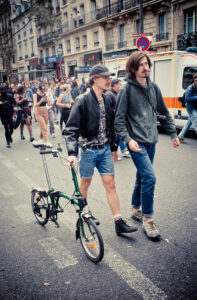 Two people walk side by side on a city street, part of a larger crowd. One pushes a small, folded green bicycle and wears a cap, leather jacket, sunglasses, and shorts. The other in a green hoodie and brown pants. Emergency vehicle visible in the background.