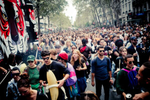 A large crowd of people gathers on a city street during a daytime event. The crowd is diverse, with individuals of various ages and styles, some holding drinks and wearing colorful clothing. Trees line the street, and promotional artwork is visible in the background.