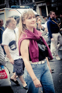 A woman with blonde hair, sunglasses perched on her head, and a maroon sweater draped over her shoulders walks through a busy street. She wears a black top, blue jeans, and carries a small bag. People and an ambulance are visible in the background.