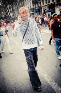 A fashionably dressed individual with light pink hair, wearing a white netted sweater and dark cargo pants, walks down a bustling city street while talking on the phone. People are seen walking and standing in the background on both sides of the street.