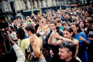 A large crowd of people at an outdoor event. Many attendees are holding up their hands, some with cameras and phones, capturing the moment. The street is bustling with activity, and buildings can be seen in the background. The atmosphere appears lively and energetic.