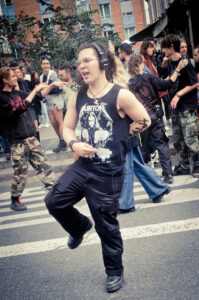 A person with headphones and a black tank top featuring a band logo passionately dances in the street amidst a group of people. The background includes a few trees and a brick building. Other individuals are chatting and moving around, creating a lively atmosphere.