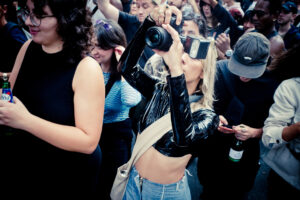 A group of people at a crowded event. A person in front, wearing a black shiny jacket and blue jeans, is holding a camera and snapping pictures. The crowd appears engaged, with individuals either looking towards the camera, holding drinks, or using their phones.