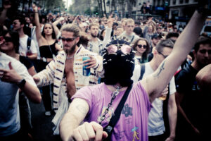 A large crowd of people is gathered outdoors, some dancing and cheering. In the foreground, a person wearing a purple shirt and a black mask with spikes and sunglasses raises their arms. Another person behind them is holding a can with raised arms. The scene is lively and energetic.