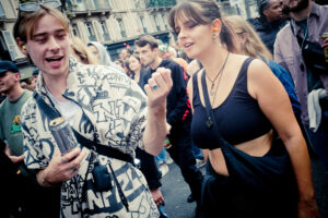 A lively crowd enjoys a street festival. In the foreground, a young man in a patterned jacket and sunglasses holds a can, talking animatedly. Beside him, a young woman in a black crop top and necklace looks engaged. People in the background are chatting and smiling.