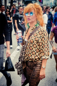A woman with red hair, wearing reflective blue sunglasses, a patterned shirt, and animal print leggings, holds a can of beer while walking in a crowd. She has a shoulder bag and large green earrings. The background includes several other people walking and buildings.