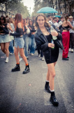20230923-DSCF0711_MOD_FRAME_resize Woman in a black dress and leather jacket holds a drink while walking along a street during a lively outdoor event. She smiles at the camera. Others in the background also enjoy the event, dressed casually in shorts, skirts, and boots, creating a festive atmosphere.
