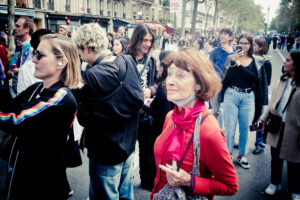 A crowd of diverse individuals, young and old, are gathered on a street. In the foreground, an older woman in a red sweater and pink scarf stands out, looking ahead. The people around her are engaged, some taking photos and videos. Buildings and trees line the background.