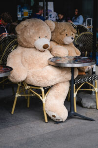 Two large nounours sit at an outdoor café table with yellow wicker chairs. One bear is significantly larger, and they both lean against the table as if they attendent their commande. People are seen in the background, indicating a busy café setting.