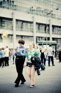 A person in a blue sheer top and black pants looks at a phone while walking beside another person in a green shirt and beige pants, carrying a drink. They are on a busy city street with more people and buildings in the background.