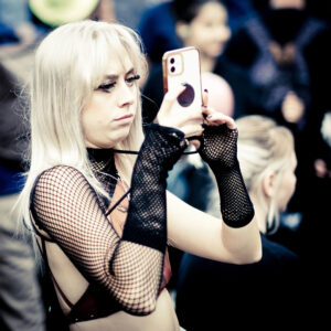 Person with long blond hair, wearing fishnet gloves and a sleeveless top, holding up a smartphone and looking attentively at it. The background is blurred, featuring a few other indistinct people.
