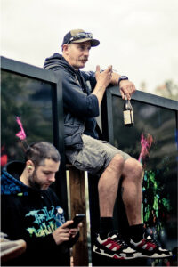 A man sits on a fence wearing a cap, sunglasses, dark hoodie, and shorts, holding a beer bottle. He looks to the side with a cigarette in one hand. Another man, with earbuds in, stands below, looking at his phone. The background features large, colorful screens.
