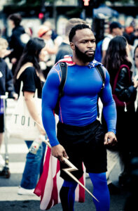 A man in a blue superhero costume with a red cape and holding a lightning bolt prop stands among a crowd of people outdoors, participating in a public event or parade.
