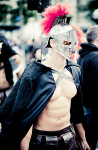 A person dressed as a Roman gladiator stands outdoors, wearing a metallic helmet with a red plume, a black cape with fur shoulders, and leather armor strapped across their chest. The background is slightly blurred with people milling about.
