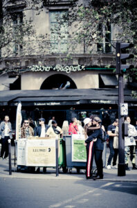 A group of people are gathered on a city sidewalk near recycling bins and signs. In the foreground, two people hug beside a traffic light, with a café and trees in the background.