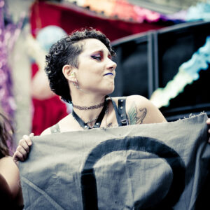 A person with short curly hair and edgy makeup, wearing a choker and a tank top, holds a banner with a partially visible symbol. The person has tattoos on their shoulder. The background is festive with colorful decorations.