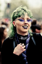 20230923-DSCF0873_MOD_FRAME_resize A person with green hair, black lipstick, and unique eyeliner smiles while wearing futuristic sunglasses, a striped tie, and a choker necklace, standing in a crowd outdoors.