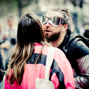 A person wearing large reflective sunglasses and a shiny jacket speaks to another person in a bright pink jacket at an outdoor event. The focus is on their interaction, while the background is blurred.